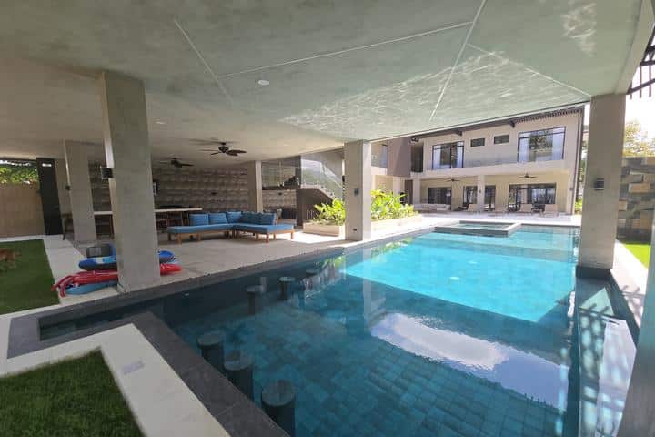 Covered pool patio with blue sectional seating and a rectangular blue-tiled pool in a modern backyard area.