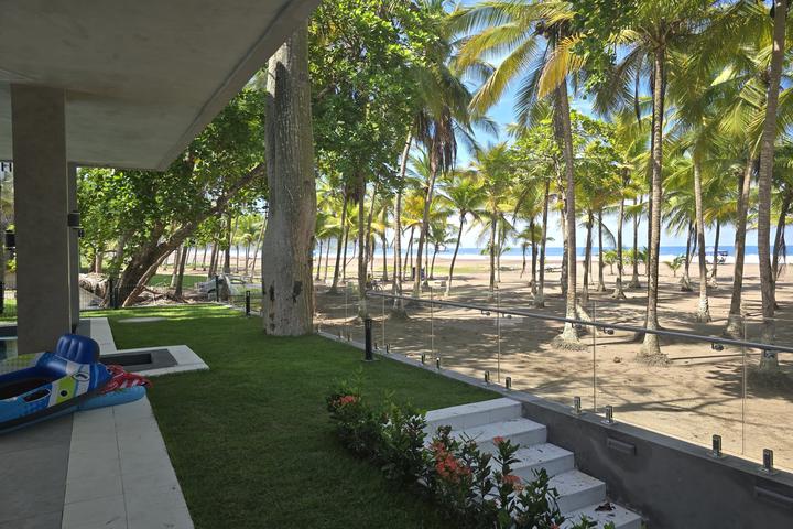Patio area overlooking a sandy beach with tall palm trees, a glass railing, and colorful inflatable toys on the left.