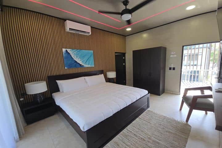 Modern bedroom with a dark wood platform bed, white bedding, and matching nightstands against a wooden slat accent wall. A ceiling fan with red LED accents, an overhead AC unit, and a large dark wardrobe are visible; a painting hangs above the bed.