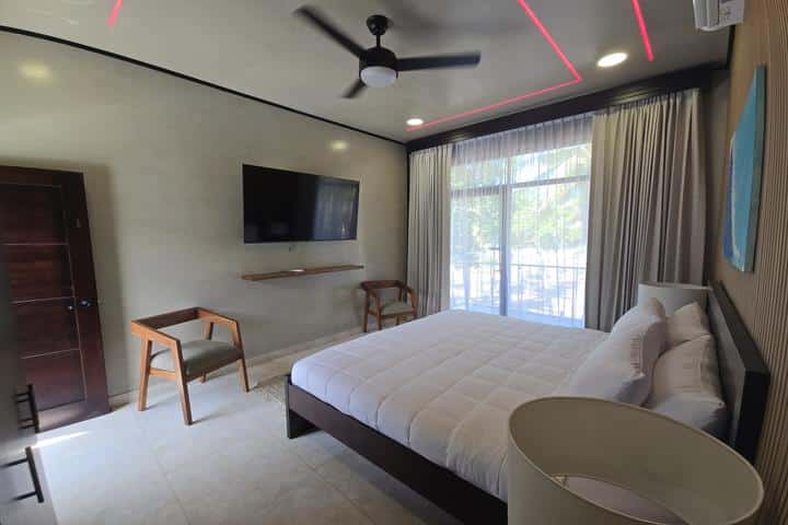Hotel bedroom with a king-size bed, ceiling fan, wall-mounted TV, two wooden chairs, and a sliding glass door to a balcony with curtains.