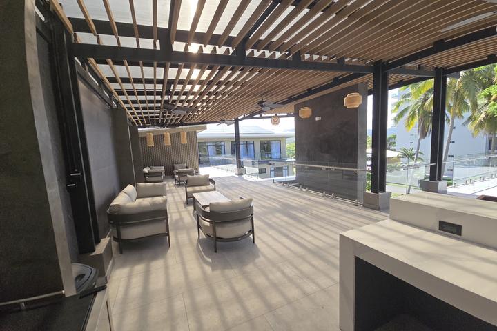 Open-air rooftop lounge with white cushioned seating under a wooden slatted pergola, glass railing, and palm trees.