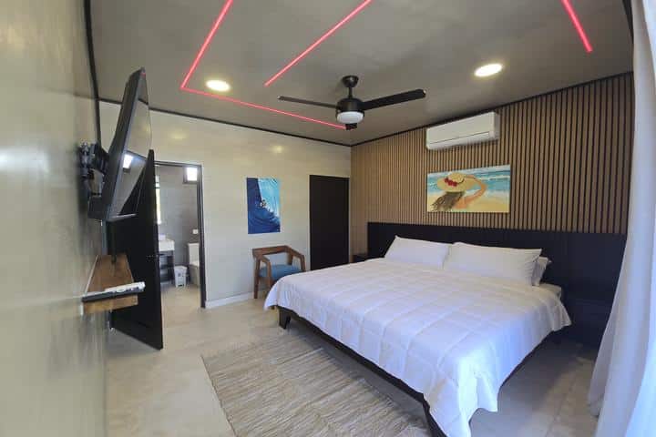 Hotel room with a king bed in white linens, dark headboard, wood slat wall, ceiling fan, and red LED light strips above. A wall-mounted TV and doorway to a bathroom are visible.