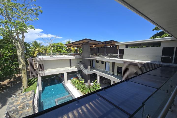 Modern hotel atrium with a rectangular swimming pool, multiple balconies, and shaded lounge areas under a central pergola, sunny blue sky above.