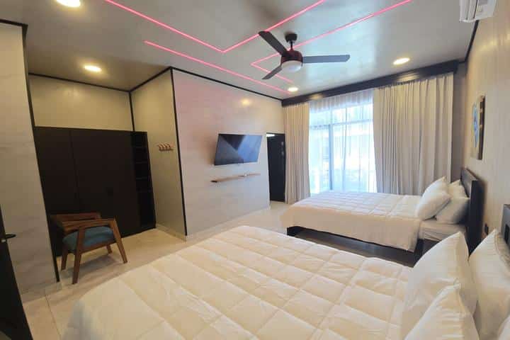 Hotel room with two double beds, wall-mounted TV, ceiling fan, and large window with curtains.