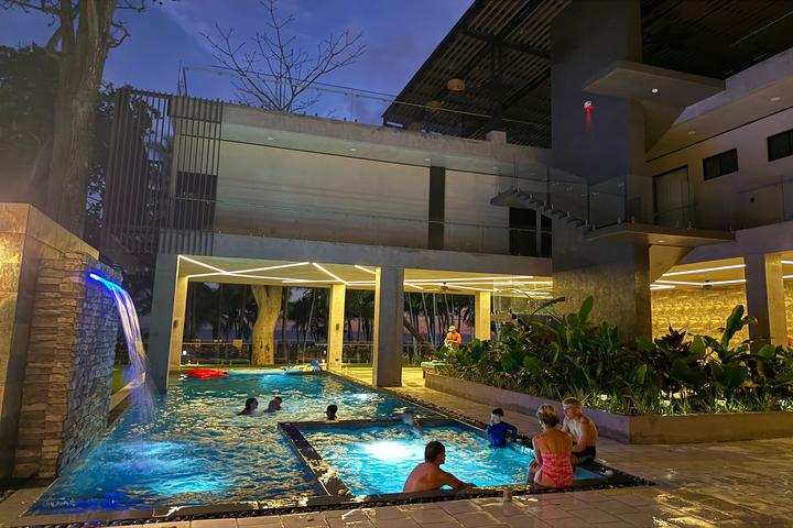 Indoor hotel pool at dusk with swimmers and a waterfall feature from a stone wall on the left.