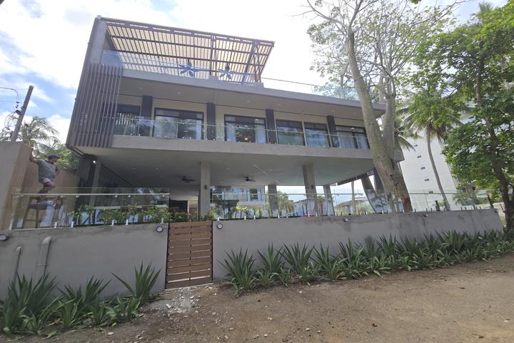 Modern two-story house with glass railings, a large balcony and elevated patio, gated by a wooden gate; low wall and row of spiky plants along the front, with a person near the gate.