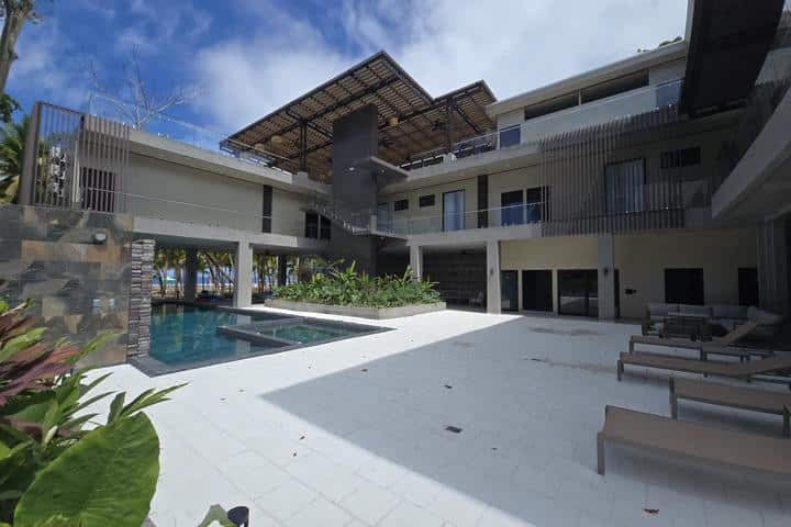 Modern hotel courtyard with a rectangular pool, sun loungers, and a shaded upper walkway around a white stone deck in a contemporary resort.