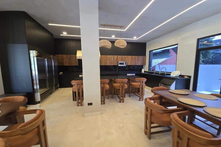 Open-plan kitchen and dining area with a central pillar, wooden tables and chairs, and plate settings on a foreground table; stainless steel appliances, dark cabinetry, and modern linear ceiling lights with large windows to the right.