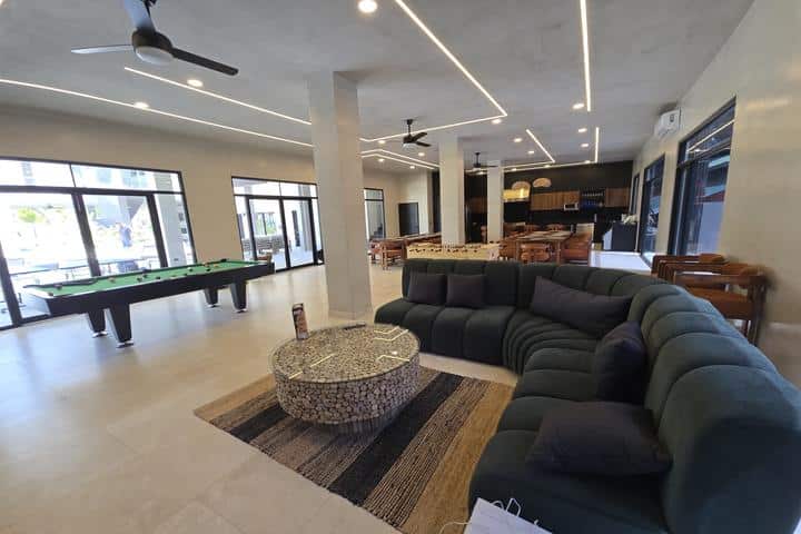 Modern recreation room with a dark green sectional sofa, a round mosaic coffee table, and a striped rug; pool tables and large windows in the background.