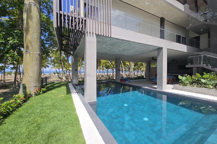 Modern beachfront pool area under a concrete building with glass railings; beach and palm trees visible beyond.