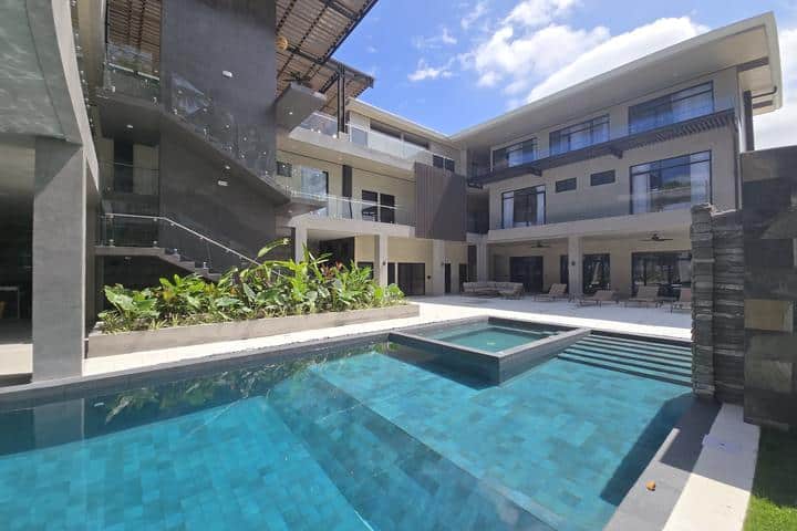 Modern multi-story hotel courtyard with a rectangular swimming pool and lounge area under a blue sky.