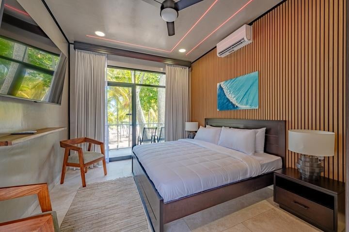 Beachfront Jaco, Costa Rica - Vacation Rental Casa Las Palmas • Costa Rica Elite Modern hotel room with a king bed, white bedding, and a wooden accent wall with a balcony view of palm trees outside.