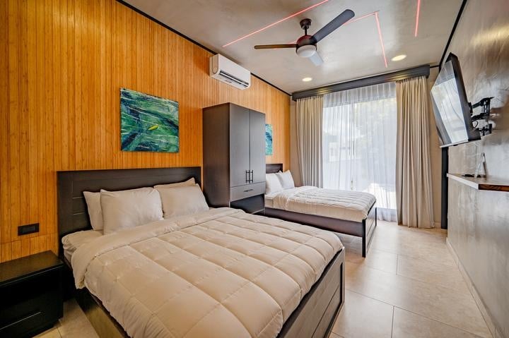 Beachfront Jaco, Costa Rica - Vacation Rental Casa Las Palmas • Costa Rica Elite Two twin beds with white comforters in a wood-paneled hotel room, plus a tall dark wardrobe, wall art, and a balcony window with sheer curtains.