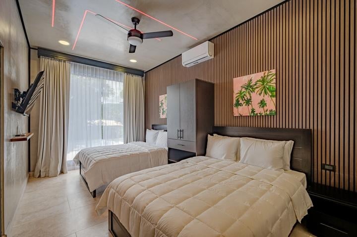 Beachfront Jaco, Costa Rica - Vacation Rental Casa Las Palmas • Costa Rica Elite Hotel room with two double beds, wood-paneled wall, and palm tree wall art; large window with sheer curtains and ceiling fan above.