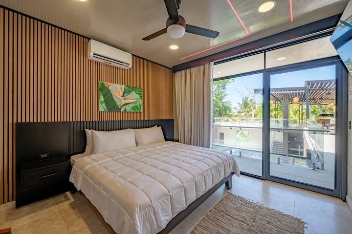 Beachfront Jaco, Costa Rica - Vacation Rental Casa Las Palmas • Costa Rica Elite Modern bedroom with a large white bed, dark headboard, and wooden slat wall; sliding glass door opens to a balcony.