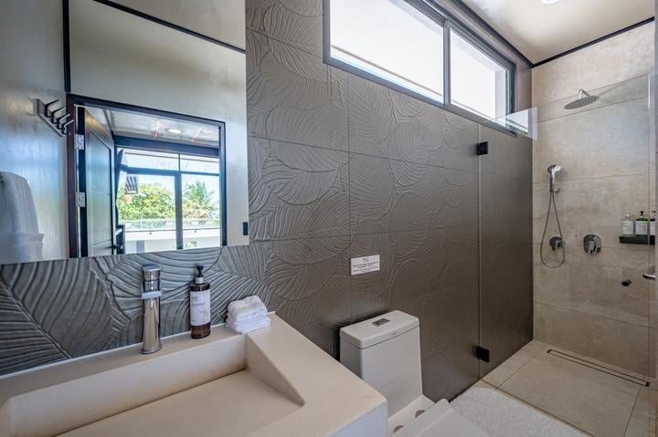 Beachfront Jaco, Costa Rica - Vacation Rental Casa Las Palmas • Costa Rica Elite Modern bathroom with textured gray wall tiles, white vanity and mirror, toilet, and a glass-enclosed walk-in shower with a rainfall showerhead.