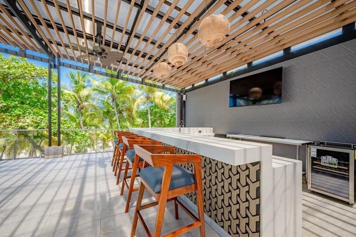 Beachfront Jaco, Costa Rica - Vacation Rental Casa Las Palmas • Costa Rica Elite Outdoor bar area with white counter, wooden bar stools with blue cushions, hanging wicker lamps, and a tropical view of palm trees outdoors