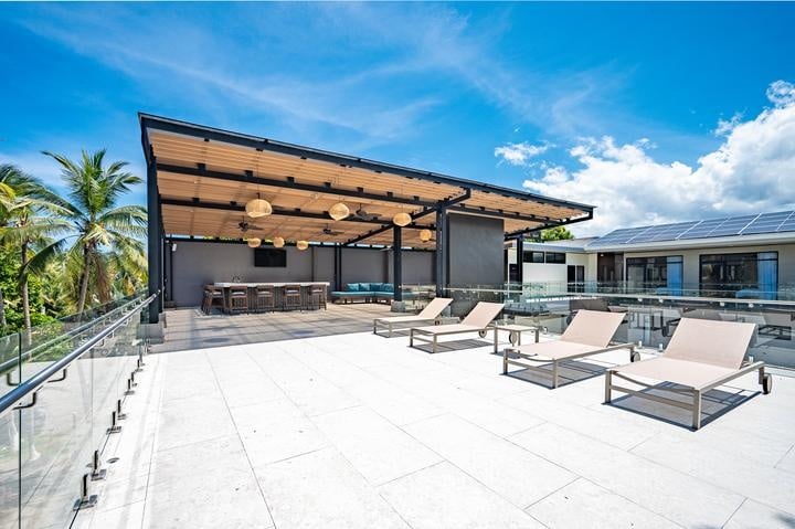 Beachfront Jaco, Costa Rica - Vacation Rental Casa Las Palmas • Costa Rica Elite Rooftop terrace with beige lounge chairs, glass railing, a shaded bar area, and palm trees under a blue sky.