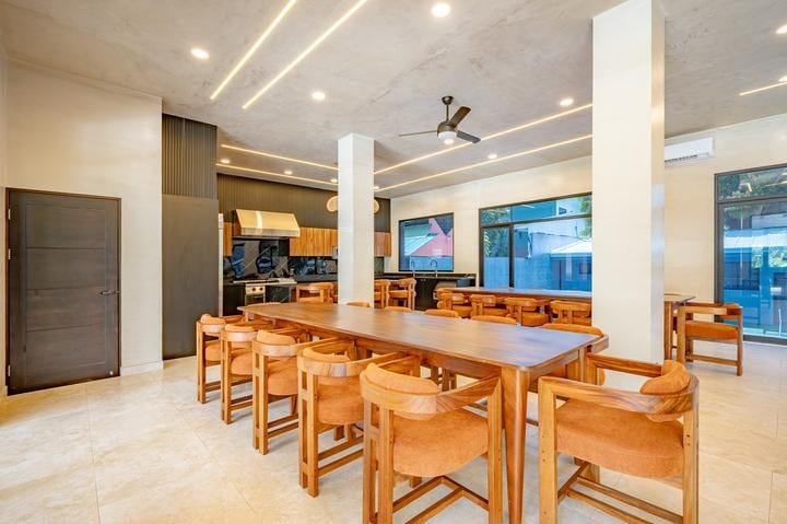 Beachfront Jaco, Costa Rica - Vacation Rental Casa Las Palmas • Costa Rica Elite Open-concept dining area with a long wooden table and many chairs, a modern kitchen in the back, and large windows letting in natural light.