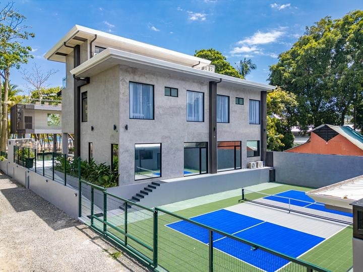 Beachfront Jaco, Costa Rica - Vacation Rental Casa Las Palmas • Costa Rica Elite Modern gray two‑story house with large windows and a flat overhang, next to a fenced blue tennis/paddle court on a sunny day.
