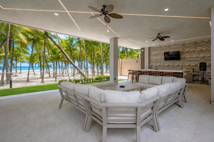 Beachfront Jaco, Costa Rica - Vacation Rental Casa Las Palmas • Costa Rica Elite Covered beachfront lounge with a white sectional sofa, ceiling fans, and a bar area overlooking palm trees and the ocean.