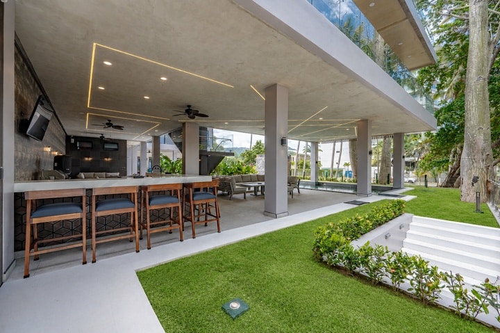 Beachfront Jaco, Costa Rica - Vacation Rental Casa Las Palmas • Costa Rica Elite Outdoor modern bar area under a covered patio with wooden stools, a long counter, and ceiling lights or fans in the ceilingled area into a lounge setting.
