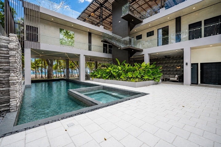 Beachfront Jaco, Costa Rica - Vacation Rental Casa Las Palmas • Costa Rica Elite Modern hotel courtyard featuring a rectangular pool with an elevated hot tub, tropical plants, and staircases to upper floors.