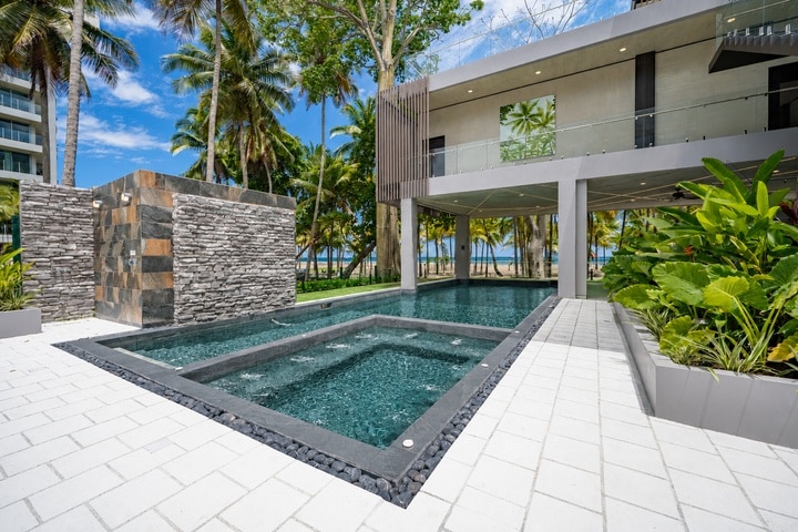 Beachfront Jaco, Costa Rica - Vacation Rental Casa Las Palmas • Costa Rica Elite Modern outdoor pool and hot tub area at a beachfront resort, with palm trees and a contemporary building in the background