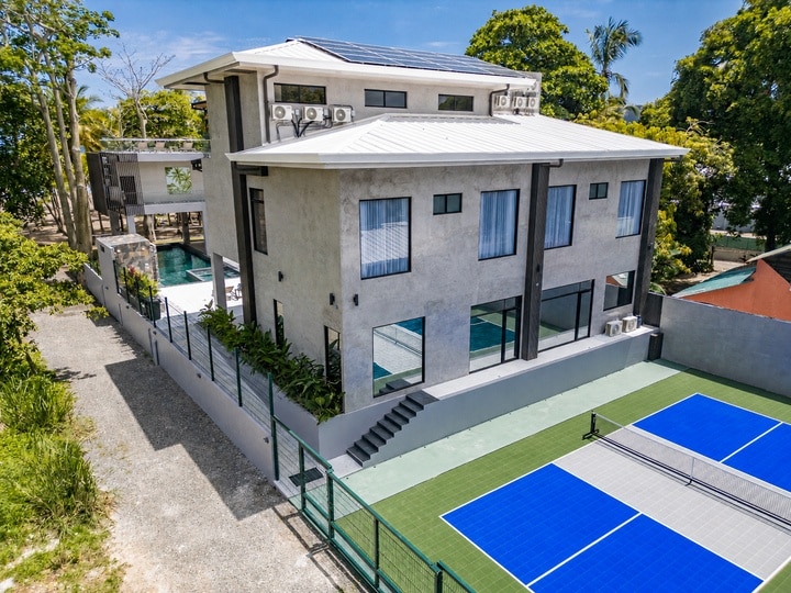 Beachfront Jaco, Costa Rica - Vacation Rental Casa Las Palmas • Costa Rica Elite A modern two-story gray house with large windows, a fenced pool area, and a blue tennis court adjacent to the yard from an elevated angle.