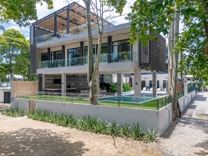 Beachfront Jaco, Costa Rica - Vacation Rental Casa Las Palmas • Costa Rica Elite Modern two-story house with glass railings, a ground-level outdoor pool, and a shaded patio area surrounded by trees.