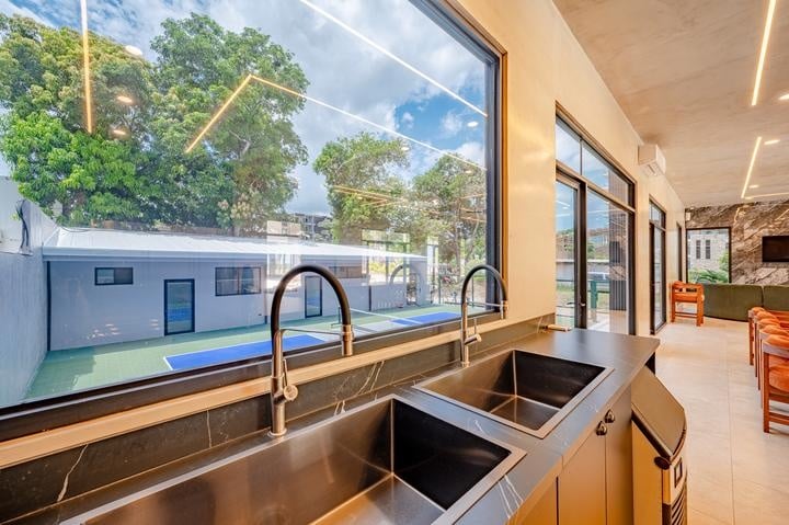 Beachfront Jaco, Costa Rica - Vacation Rental Casa Las Palmas • Costa Rica Elite Double stainless steel kitchen sink with two faucets, facing a large window overlooking an outdoor tennis court and trees outside.