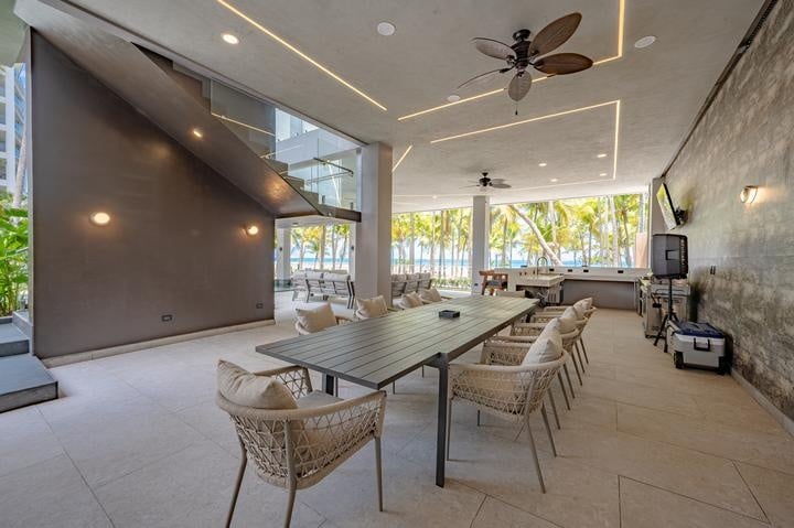 Beachfront Jaco, Costa Rica - Vacation Rental Casa Las Palmas • Costa Rica Elite Indoor lounge with a long dining table, woven chairs, and ceiling fans, opening to a tropical palm view through large windows in natural light.