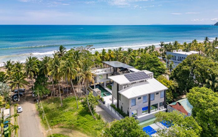 Aerial view of a modern beachfront hotel with solar panels on the roof, palm trees, and a blue ocean backdrop.